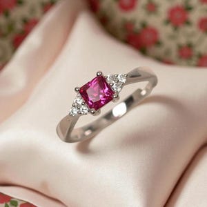 May include: A silver ring with a striking square, pink gemstone at its centre, complemented by smaller, clear stones. The ring is displayed on a soft, pink fabric, creating a refined look. This piece of jewellery is suitable for special events and celebrations.