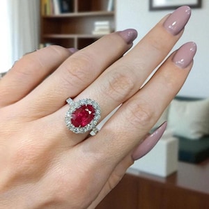 Könnte beinhalten: Ein silberfarbener Ring mit einem großen, oval geschliffenen, leuchtend roten Edelstein in der Mitte, der von einem Kranz aus kleinen, klaren, rund geschliffenen weißen Steinen umgeben ist. Die Ringschiene ist schmal und metallisch gehalten.