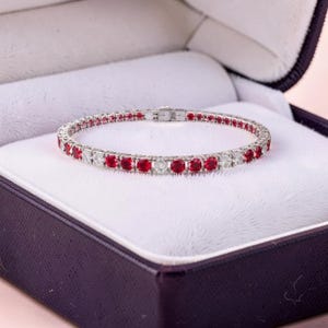 May include: A silver bracelet with alternating red and clear gemstones, displayed in a dark purple jewelry box with a white interior. The bracelet is a tennis style, with a clasp closure.
