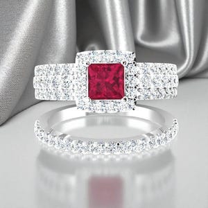 May include: A two-piece ring set featuring a square, deep red gemstone center, surrounded by a halo of smaller, clear stones. The band is also adorned with clear stones, creating a sparkling effect. The rings are set on a reflective surface.