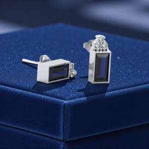 Pode incluir: Um par de brincos de tarracha com uma safira azul escura retangular de corte baguette engastada numa armação de metal prateado, acentuada com um grupo de três pequenas pedras redondas transparentes no topo e um fecho de segurança.