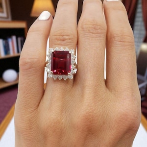 Könnte beinhalten: Ein goldener Ring mit einem großen, rechteckigen, tiefroten Edelstein, umgeben von einem Kranz aus kleineren, klaren Steinen. Der Ring wird an einer Hand mit weißem Nagellack präsentiert, wodurch das Design und die Farbe des Schmucks hervorgehoben werden.