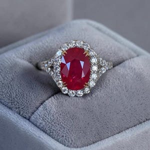 Peut inclure: Une bague avec un rubis rouge profond taille ovale serti sur une monture en métal argenté. Le rubis central est entouré d'un halo de petits diamants blancs ronds et clairs. L'anneau présente des diamants supplémentaires sur les côtés.
