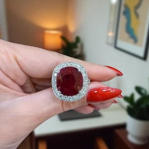 May include: A large, silver ring featuring a vibrant, oval-cut ruby gemstone. The ruby is surrounded by a halo of smaller, clear gemstones. The ring is held up against a blurred background.