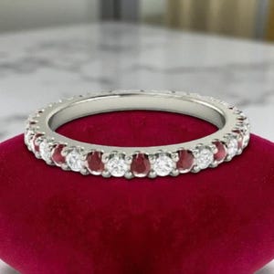 May include: A silver ring featuring alternating round white and red gemstones. The ring is displayed on a red velvet surface, highlighting the sparkle of the gems. The ring is a full eternity band, with gemstones encircling the entire band.