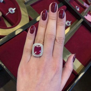 May include: A ring featuring a large, rectangular, deep red gemstone set in a silver-toned metal band. The central stone is surrounded by a double halo of small, clear, sparkling stones. The ring is worn on a finger with dark red nail polish.
