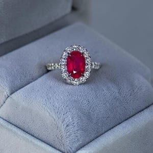 Peut inclure: Une bague ornée d'un rubis rouge vif de taille ovale au centre, entouré d'un halo de petits diamants blancs ronds et transparents. L'anneau est en métal argenté poli et comporte des diamants supplémentaires le long de la tige.
