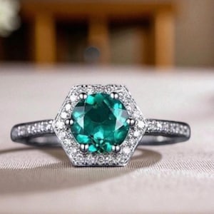 May include: A silver ring featuring a hexagonal halo of small, clear stones encircling a large, round, emerald-green gemstone. The ring band is also set with small, clear stones. The ring is displayed against a neutral backdrop.