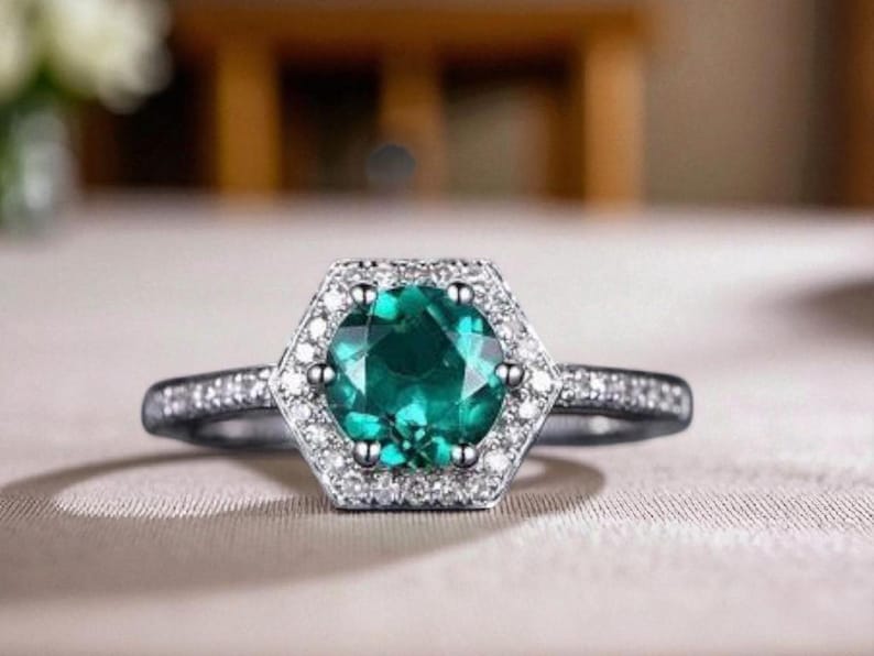 May include: A silver ring featuring a hexagonal halo of small, clear stones encircling a large, round, emerald-green gemstone. The ring band is also set with small, clear stones. The ring is displayed against a neutral backdrop.