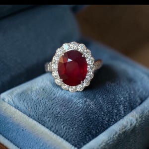Puede incluir: Un anillo con una gran piedra preciosa de color rojo intenso y corte ovalado en el centro, rodeada por un halo de pequeñas piedras blancas claras y redondas. El engaste es metálico y el anillo se presenta en un estuche de terciopelo azul.