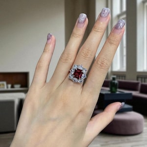 May include: A silver-toned ring featuring a large, rectangular, deep red gemstone at the center, surrounded by a halo of smaller, round, clear white stones arranged in a floral-like pattern on a polished metal band.
