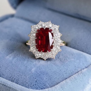 Può includere: Un anello d'oro con una grande gemma rosso scuro a taglio ovale, circondata da un alone di gemme più piccole e chiare. L'anello è posto su uno sfondo di velluto blu, che suggerisce un portagioie.