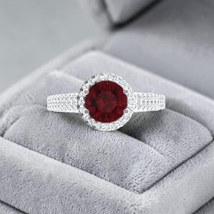 Peut inclure: Une bague ornée d'une pierre précieuse ronde rouge foncé en son centre, entourée d'un halo de petites pierres claires et scintillantes. L'anneau est en métal et est incrusté de plusieurs rangées de minuscules pierres claires taillées.