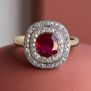 Peut inclure: Une bague en or avec une grosse pierre centrale rouge rubis, entourée de deux rangées de petits diamants clairs. Le corps de la bague est en or.
