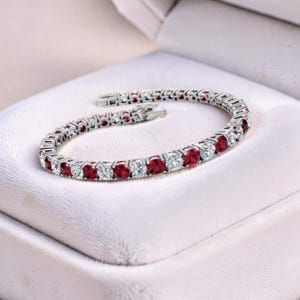 May include: A silver bracelet with alternating red and clear gemstones. The bracelet is displayed open inside a white jewelry box. The gemstones are round and set closely together, creating a continuous line of sparkle.