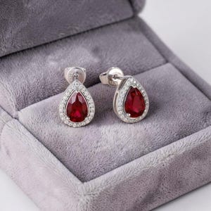 May include: A pair of silver-toned stud earrings featuring a pear-shaped red gemstone centre surrounded by a halo of small clear stones. The metal settings are marked with S925, and the earrings are presented in a soft, grey velvet-lined box.