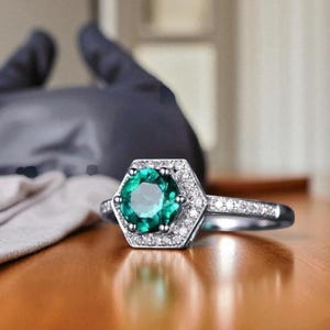 May include: A silver ring featuring a hexagonal emerald-cut green gemstone, encircled by small, clear accent stones. The ring is displayed on a wooden surface. The ring's band is also adorned with small stones.
