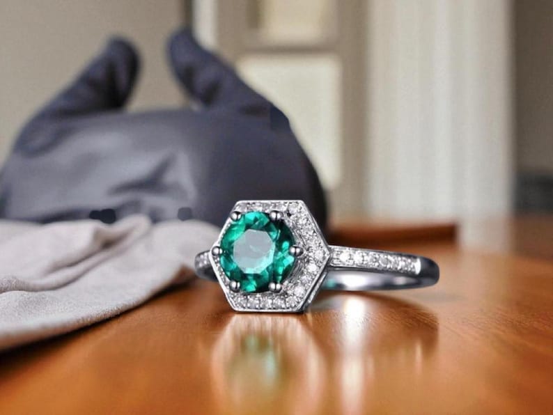 May include: A silver ring featuring a hexagonal emerald-cut green gemstone, encircled by small, clear accent stones. The ring is displayed on a wooden surface. The ring's band is also adorned with small stones.