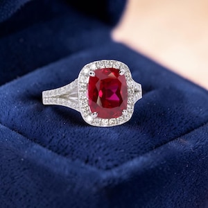 Könnte beinhalten: Ein Ring mit einem großen, tiefroten Edelstein im Kissenschliff in der Mitte, umgeben von einem Kranz aus kleinen, klaren, rund geschliffenen Steinen. Die Ringschiene ist gespalten und mit weiteren kleinen, klaren Steinen besetzt.
