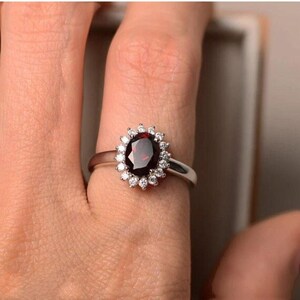 Könnte beinhalten: Ein silberfarbener Ring mit einem zentralen, oval geschliffenen, tiefroten Edelstein, der von einem Kranz aus kleinen, klaren, rund geschliffenen Steinen umgeben ist. Die Ringschiene ist glatt und poliert und bildet einen Kontrast zum roten Stein.