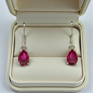 Peut inclure: Une paire de boucles d'oreilles pendantes avec des pierres précieuses rouges en forme de poire. Les boucles d'oreilles sont serties dans un métal argenté et présentées dans un écrin crème ouvert. Les pierres ont une taille à facettes.