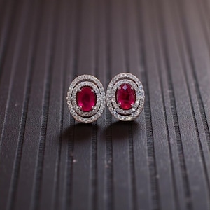 May include: A pair of stud earrings featuring oval-shaped, deep red gemstones surrounded by a halo of smaller, clear stones. The earrings are set in a silver-colored metal and rest on a dark, textured surface.