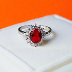 Peut inclure: Une bague argentée ornée d'une grande pierre centrale rouge vif de taille ovale. La pierre précieuse rouge est entourée d'un halo de petites pierres blanches claires et étincelantes montées sur griffes, formant un motif floral classique.