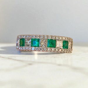 Może przedstawiać: Pierścionek z różowego złota z rzędem kwadratowych szmaragdów i diamentów. Szmaragdy są w żywym zielonym kolorze, a diamenty błyszczą. Pierścionek jest osadzony małymi, okrągłymi diamentami wokół szmaragdów i diamentów.