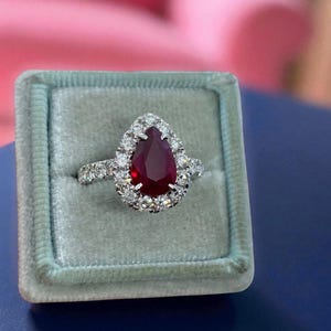 Op de afbeelding: Een ring met een peervormige rode robijn in een zilverkleurige metalen band. De centrale robijn is omringd door een halo van kleine, ronde, heldere witte stenen, met extra heldere stenen ingelegd langs de zijkanten van de ringband.
