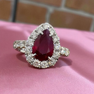 May include: A pear-shaped ruby ring with a halo of small diamonds. The ring's band is also encrusted with diamonds, and the setting is silver-toned. The ruby is a deep red color, and the ring is set against a pink background.