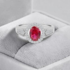 Peut inclure: Une bague argentée ornée d'une pierre rouge centrale de taille ovale, entourée d'un halo de petites pierres blanches. L'anneau présente des pierres blanches supplémentaires incrustées sur les côtés de la monture centrale ovale.