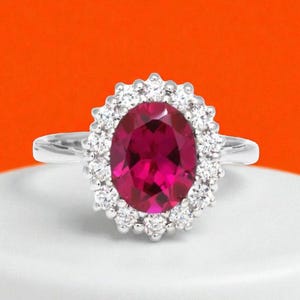 Peut inclure: Une bague argentée ornée d'une grande pierre précieuse ovale rose foncé au centre. La pierre centrale est entourée d'un halo de petites pierres claires rondes serties dans une monture en métal festonnée, sur un anneau argenté poli.