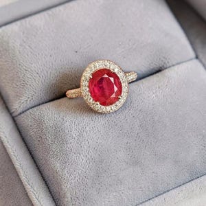 Può includere: Un anello in oro rosa con una gemma rossa vibrante dal taglio ovale al centro, circondata da un alone di piccoli diamanti bianchi incastonati a pavé. Il gambo dell'anello è sottile e anch'esso tempestato di minuscoli diamanti bianchi.