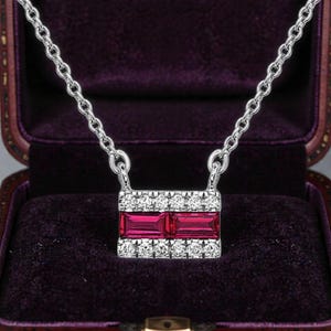 May include: Silver necklace with a rectangular pendant featuring two rectangular, deep red gemstones. The pendant is bordered with small, clear crystals. The necklace is displayed inside a purple jewelry box.