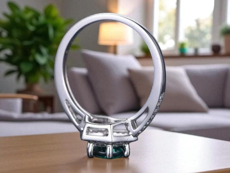 May include: A silver ring featuring a square, emerald-cut green gemstone. The ring band is embellished with small, clear stones. The ring is set on a wooden surface, with a blurred living room background.