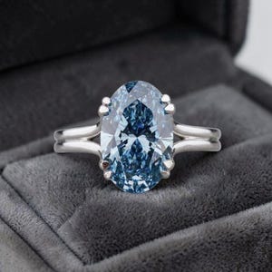Puede incluir: Un anillo con una gran piedra preciosa de color azul claro y corte ovalado, montada en una banda de metal pulido de tono plateado. La piedra está asegurada por cuatro garras y la banda presenta un diseño de brazo dividido en ambos lados.