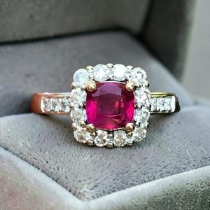 Könnte beinhalten: Ein Ring mit einem zentralen, kissenförmigen, tiefroten Rubin, der von einem Kranz aus kleinen, runden, weißen Diamanten umgeben ist. Die Ringschiene besteht aus einem metallischen Material und ist seitlich mit weiteren kleinen weißen Diamanten besetzt.