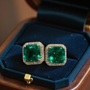 May include: A pair of square-cut emerald earrings set in gold with a halo of small, clear gemstones. The earrings are displayed in a dark blue velvet jewelry box with a wooden exterior.