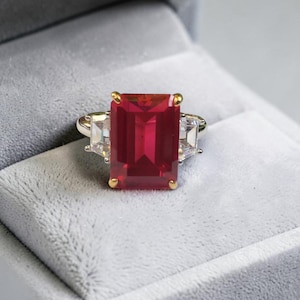 Op de afbeelding: Een ring met een grote, rechthoekige, smaragdgeslepen rode edelsteen gezet in gouden klauwen. De middelste steen wordt geflankeerd door twee kleinere, heldere, trapeziumvormige stenen aan elke kant. De ringband is gemaakt van gepolijst zilverkleurig metaal.