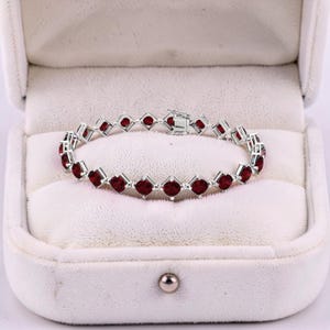 May include: A silver bracelet with square-cut, deep red gemstones. The bracelet is displayed inside a white, velvet-lined jewelry box. The gemstones are evenly spaced and set in a silver metal. The clasp is visible, and the bracelet is centered in the box.