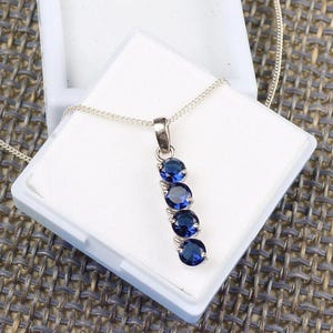 May include: A silver pendant necklace featuring four round, deep blue gemstones set in a vertical line. The necklace is displayed in a white jewelry box, with the chain visible. The gemstones have a vibrant, polished appearance.