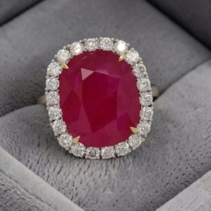 Op de afbeelding: Een ring met een grote, ovaal geslepen, dieprode edelsteen als middelpunt, omringd door een halo van kleine, ronde, briljant geslepen witte diamanten. De centrale steen wordt vastgehouden door vier geelgouden klauwen in een witmetalen zetting.