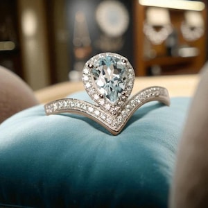 Puede incluir: Anillo plateado con una piedra preciosa azul claro en forma de pera en el centro, rodeada de piedras más pequeñas y transparentes. La banda forma una V, también engastada con pequeñas piedras transparentes. El anillo descansa sobre una superficie de terciopelo azul.