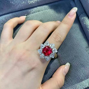 Könnte beinhalten: Ein silberfarbener Ring mit einem großen, quadratisch geschliffenen, leuchtend roten Edelstein in der Mitte, umgeben von einem Kranz aus acht birnenförmigen, klaren weißen Steinen in Blütenform auf einer polierten Metallschiene.