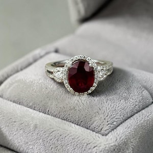 Puede incluir: Un anillo de tono plateado con una gran piedra preciosa de color rojo intenso y corte ovalado en el centro. La piedra central está rodeada por un halo de pequeñas piedras claras de corte redondo. La banda también tiene pequeñas piedras incrustadas.
