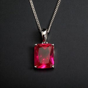 Pode incluir: Um colar de prata com um pingente retangular vermelho rubi com corte radiante. O pingente é cravejado em uma moldura de prata e pendurado em uma corrente delicada. A pedra preciosa é de uma cor vermelha profunda e vibrante.