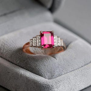 May include: A ring featuring a rectangular emerald-cut vibrant pink center stone flanked by three small clear stones on each side, all set in a polished metallic band. The design showcases a stepped arrangement of the side stones leading to the center.