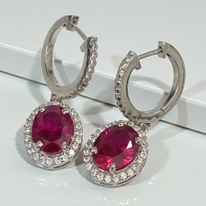 May include: A pair of silver hoop earrings. Each earring features an oval, deep red gemstone, encircled by smaller, clear stones. The earrings are presented on a white surface, highlighting their elegant design and vibrant colour.