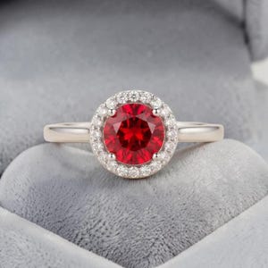 May include: A ring featuring a round, vibrant red centre stone surrounded by a halo of small, clear, sparkling stones. The band is a smooth, polished silver-toned metal, and the setting is designed with four prongs securing the centre stone.