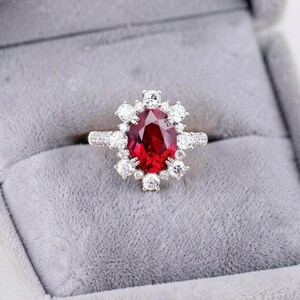 Puede incluir: Un anillo de tono plateado con una gran piedra preciosa de color rojo intenso y corte ovalado en el centro. La piedra central está rodeada por un halo de doce piedras blancas claras más pequeñas de corte redondo, con piedras adicionales.